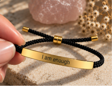 Load image into Gallery viewer, A hand holds a black braided cord bracelet with a polished gold ID plate engraved with the words 'I am enough'. The background features a pink crystal and dried white flowers on a textured beige surface.
