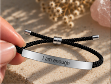 Load image into Gallery viewer, A hand holds a black braided cord bracelet with a polished silver metal ID plate engraved with the words 'I am enough'. The background is a warm, sandy beige surface with soft-focus dried flowers and a pink crystal.
