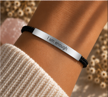 Load image into Gallery viewer, A close-up of a person's wrist wearing a silver bar bracelet engraved with the words 'I am enough', paired with a black cord bracelet and a cream knit sleeve, set against a soft-focus background of dried flowers and a pink stone.

