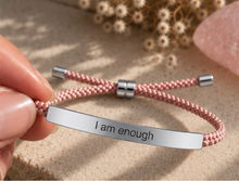 Load image into Gallery viewer, A close-up of a hand holding a pink braided cord bracelet with a silver metal bar engraved with the words 'I am enough'. The background is a textured beige surface with dried flowers and a pink stone.
