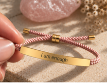 Load image into Gallery viewer, A close-up of a hand holding a pink braided cord bracelet with a gold-tone metal ID bar engraved with the words 'I am enough'. The bracelet has gold-tone sliding adjusters and end caps, and is set against a textured beige background with soft-focus decorative elements.
