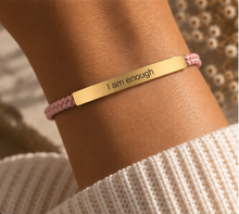 Load image into Gallery viewer, A close-up of a person's wrist wearing a gold bar bracelet engraved with the words 'I am enough' in black text, paired with a pink braided cord bracelet, set against a soft beige knit sweater.
