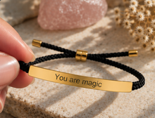 Load image into Gallery viewer, A close-up of a hand holding a black braided cord bracelet with a polished gold-tone ID bar engraved with the words 'You are magic'. The background features a textured beige stone surface with a pink rose quartz crystal and dried flowers.

