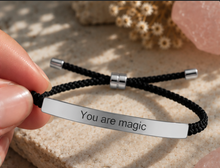 Load image into Gallery viewer, A close-up of a hand holding a black braided cord bracelet with a polished silver bar engraved with the words 'You are magic'. The background features a textured beige stone with soft-focus dried flowers and a pink crystal.
