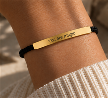 Load image into Gallery viewer, A close-up of a person's wrist wearing a black braided cord bracelet with a polished gold bar engraved with the words 'You are magic'.
