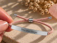 Load image into Gallery viewer, A close-up of a hand holding a pink braided cord bracelet with a polished silver bar engraved with the words 'You are Magic'. The background features a textured beige surface with dried flowers and a pink stone.
