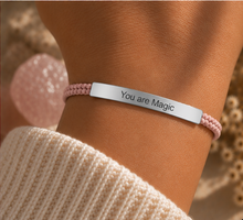 Load image into Gallery viewer, A close-up of a person's wrist wearing a pink braided cord bracelet with a polished silver metal bar engraved with the words 'You are Magic'. The person is wearing a cream-colored knit sweater, and a pink rose quartz crystal is visible in the soft-focus background.
