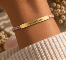Load image into Gallery viewer, A close-up of a person's wrist wearing a gold ID bracelet engraved with 'You are Magic' on a light pink braided cord bracelet, styled with a cream ribbed knit sweater.
