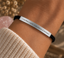 Load image into Gallery viewer, A close-up of a person's wrist wearing a black cord bracelet with a polished silver rectangular plate engraved with the words 'I manifest a world of abundance &amp; kindness'. The person is wearing a cream-colored knit sweater, and the background is a soft-focus beige and pink setting.
