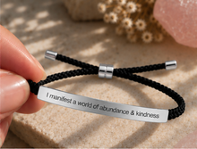 Load image into Gallery viewer, A close-up of a hand holding a black braided cord bracelet with a polished silver metal ID plate engraved with the phrase 'I manifest a world of abundance &amp; kindness'. The background consists of a textured beige stone surface with soft-focus decorative elements.
