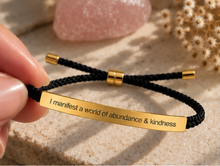 Load image into Gallery viewer, A close-up of a hand holding a black braided cord bracelet with a polished gold bar engraved with the phrase 'I manifest a world of abundance &amp; kindness'. The background features a soft-focus pink stone and dried flowers on a textured beige surface.

