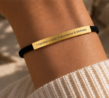 Load image into Gallery viewer, A close-up of a person's wrist wearing a black cord bracelet with a polished gold rectangular plate engraved with the quote, 'I manifest a world of abundance &amp; kindness'.
