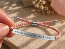 Load image into Gallery viewer, A close-up of a hand holding a pink braided cord bracelet with a silver metal ID plate engraved with the phrase 'I manifest a world of abundance &amp; kindness'. The bracelet features silver metal end caps and sliding silver metal beads.
