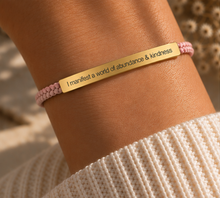 Load image into Gallery viewer, A close-up of a person's wrist wearing a gold bar bracelet engraved with the phrase 'I manifest a world of abundance &amp; kindness' on a pink braided cord bracelet, paired with a cream knit sweater.
