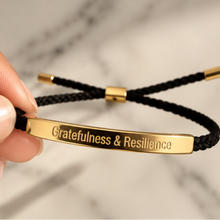 Load image into Gallery viewer, Hand holding a black cord bracelet with a gold Gratefulness and Resilience bar - Let's Jam, Camden Market London
