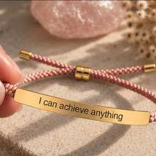 Load image into Gallery viewer, Hand holding a pink cord bracelet with a gold engraved bar beside a crystal - Let's Jam, Camden Town London
