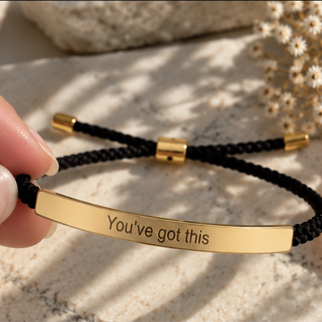 Hand holding a black cord bracelet with a gold You've Got This bar - Let's Jam jewellery, Camden Market