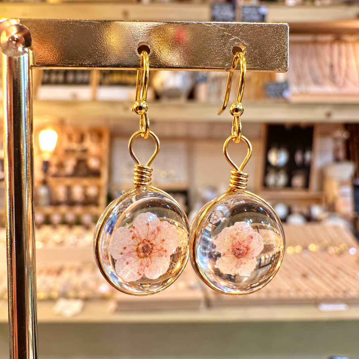 Round resin earrings with real baby pink blossoms hanging on a gold stand in the shop - Let's Jam, Camden Market, London