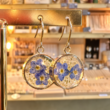Load image into Gallery viewer, Round resin earrings filled with little real blue flowers hanging on a stand in the shop - Let's Jam jewellery, Camden Town
