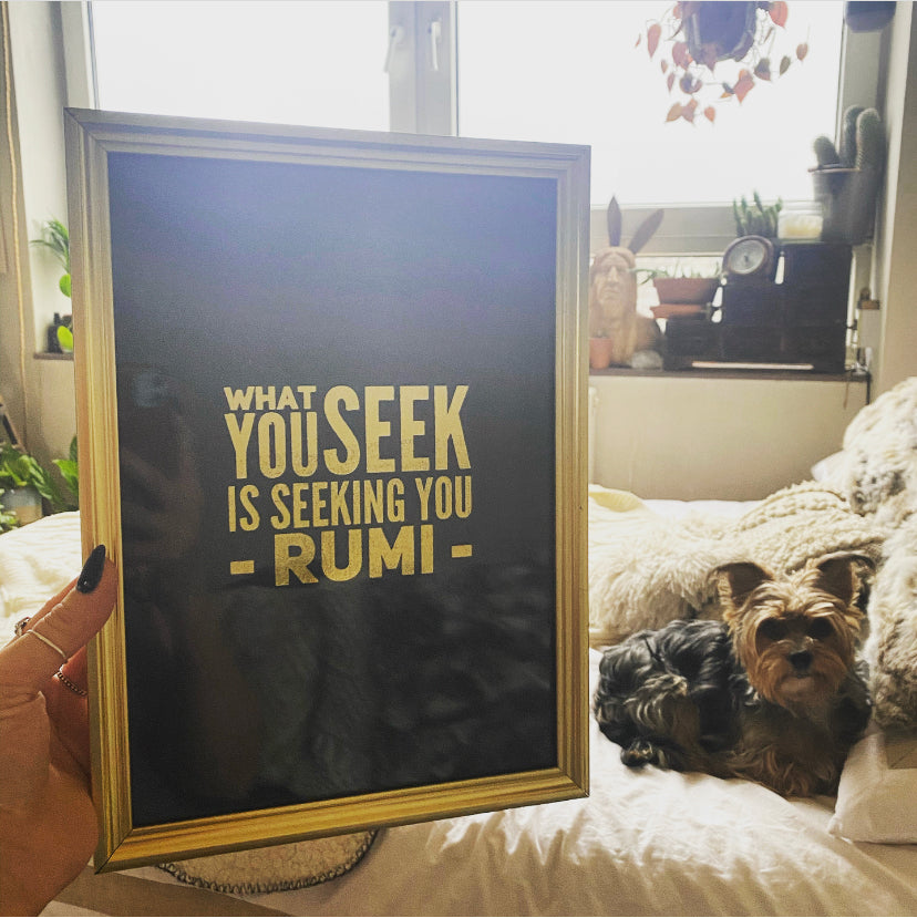 A gold-framed print of the Rumi quote "What you seek is seeking you" in gold lettering, held up beside a small dog on a bed - Let's Jam print collection