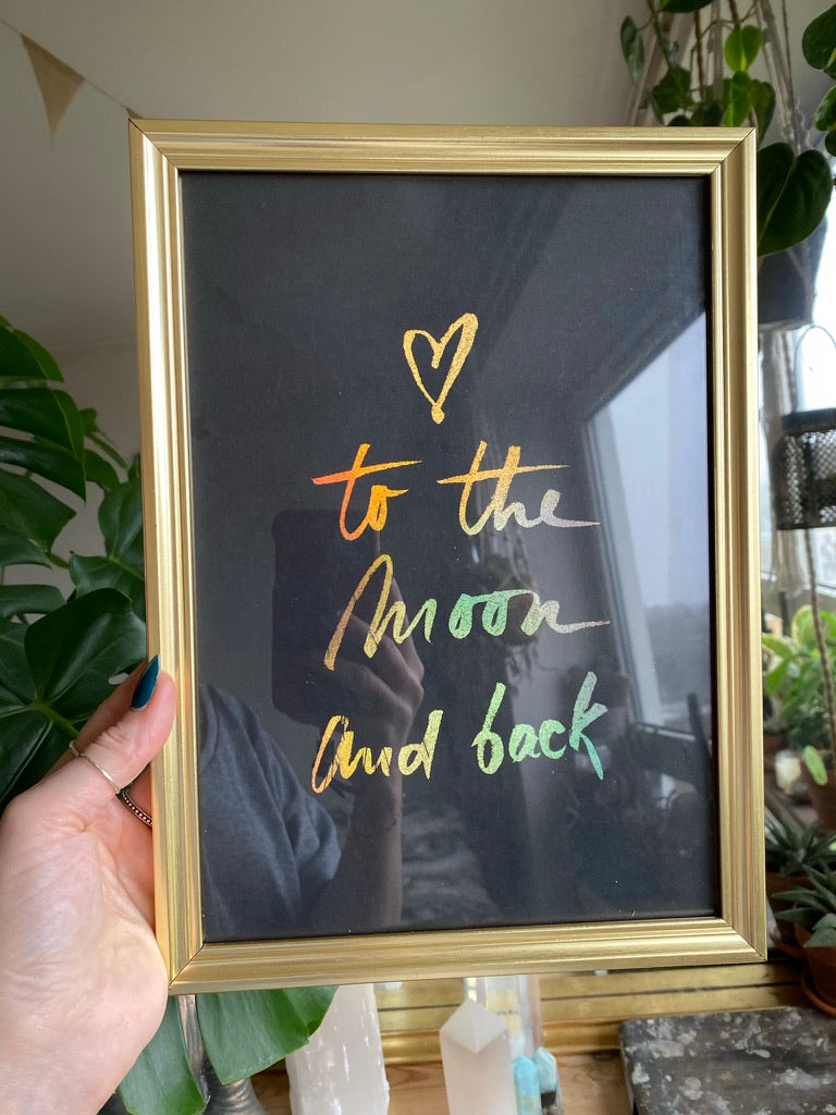 A hand holding a gold-framed 'To the Moon and Back' hand-lettered print in holographic gold and rainbow foil on black velvet paper, with a heart above the text - Let's Jam print collection