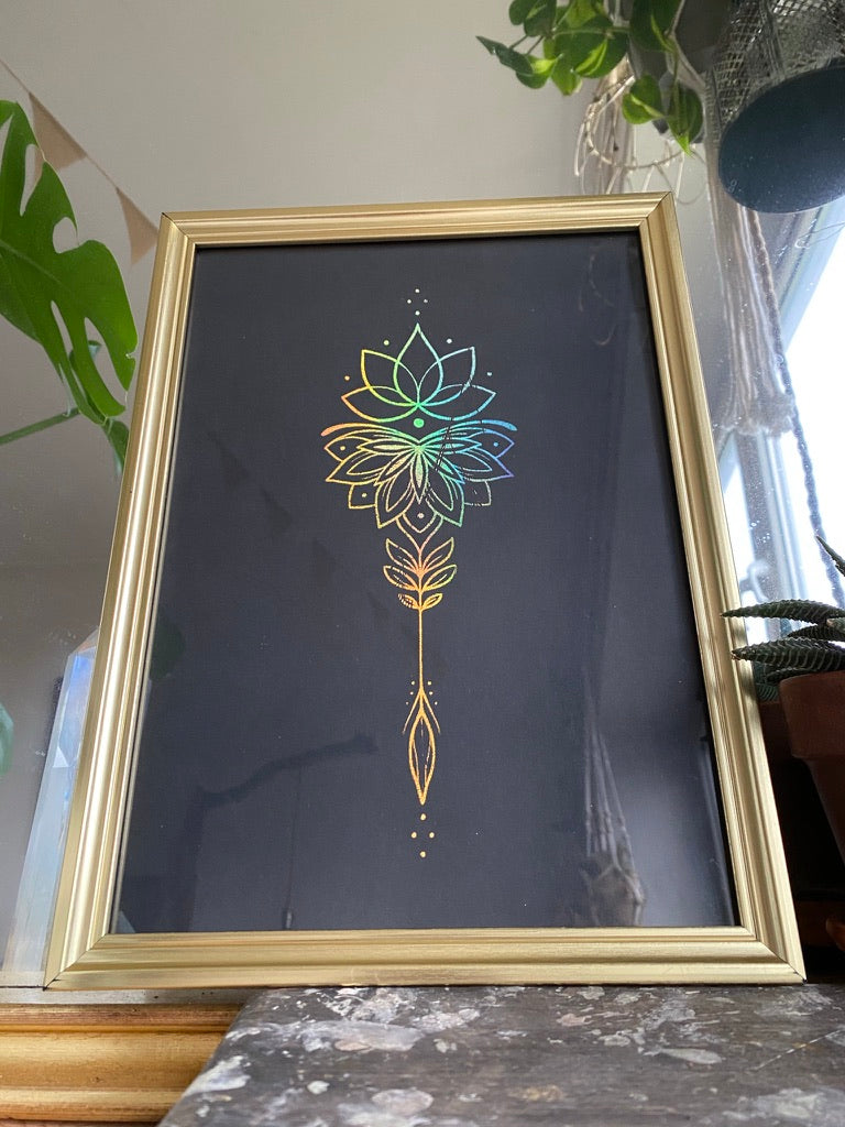 A gold-framed rainbow holographic print of an Unalome mandala symbol with a lotus flower and trailing spiral, displayed on a table beside houseplants - Let's Jam print collection