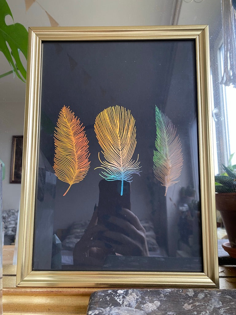 A gold-framed holographic print of three rainbow feathers in orange, yellow and blue, displayed on a table beside a houseplant - Let's Jam print collection