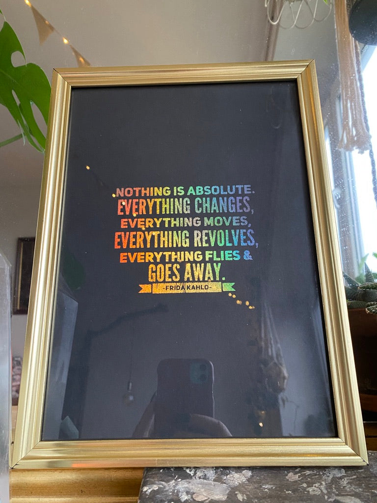 A gold-framed rainbow holographic print of a Frida Kahlo quote about nothing being absolute and everything changing, displayed against a window with fairy lights - Let's Jam print collection