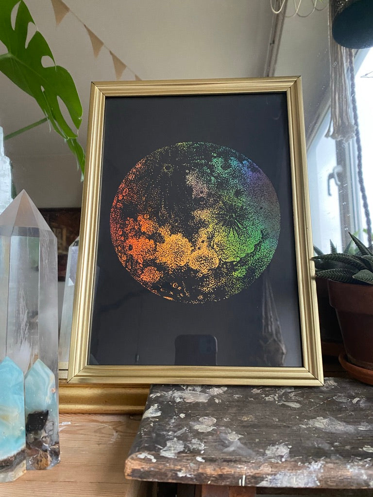 A gold-framed rainbow holographic stippled art print of the moon, displayed on a wooden table beside crystal points and a houseplant - Let's Jam print collection