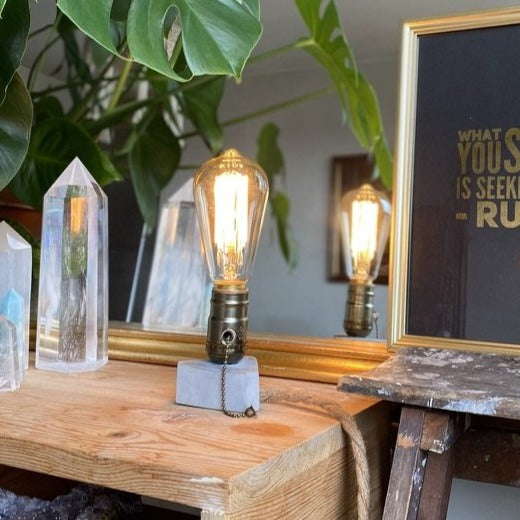 Triangle concrete lamp with a glowing Edison-style filament bulb, mirrored beside clear quartz crystal points on a wooden shelf with a framed Rumi print reflected behind - Let's Jam collection