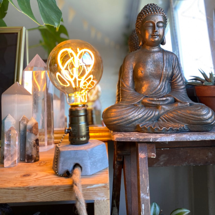 Concrete lamp with a heart-shaped LED filament bulb glowing warmly, displayed beside quartz crystal points with a bronze Buddha statue in the background - Let's Jam collection