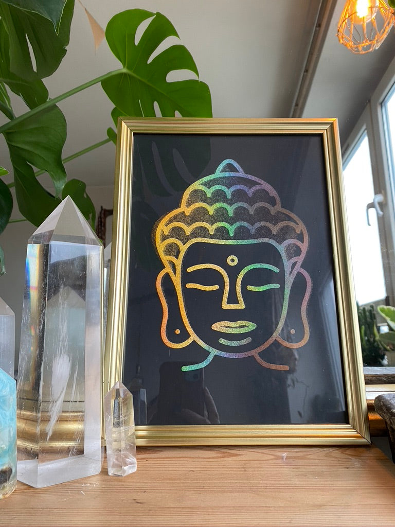 A gold-framed rainbow holographic line-art print of a Buddha head, displayed on a wooden table beside a clear quartz crystal point and a houseplant - Let's Jam print collection