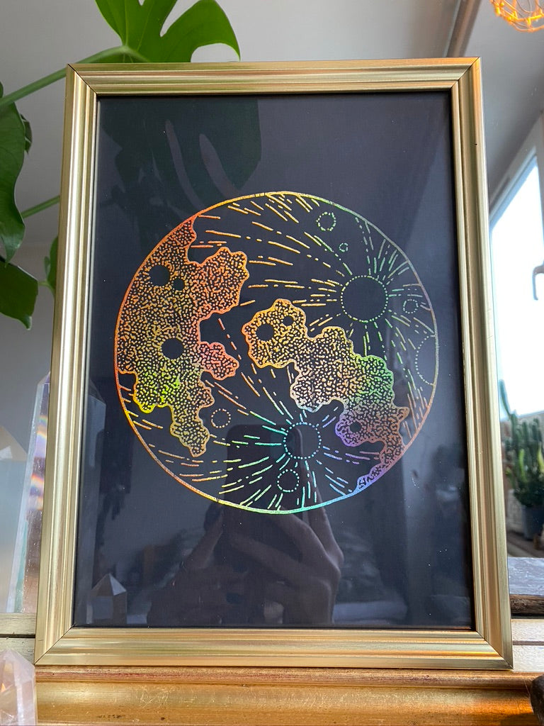 A gold-framed rainbow holographic stippled art print of a moon design with sunburst rays, displayed on a wooden table beside a houseplant - Let's Jam print collection