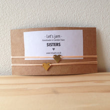 Load image into Gallery viewer, Two matching friendship bracelets with small gold heart charms, displayed on a kraft card that reads 'Sisters', perfect for gifting a sibling - Let's Jam jewellery collection
