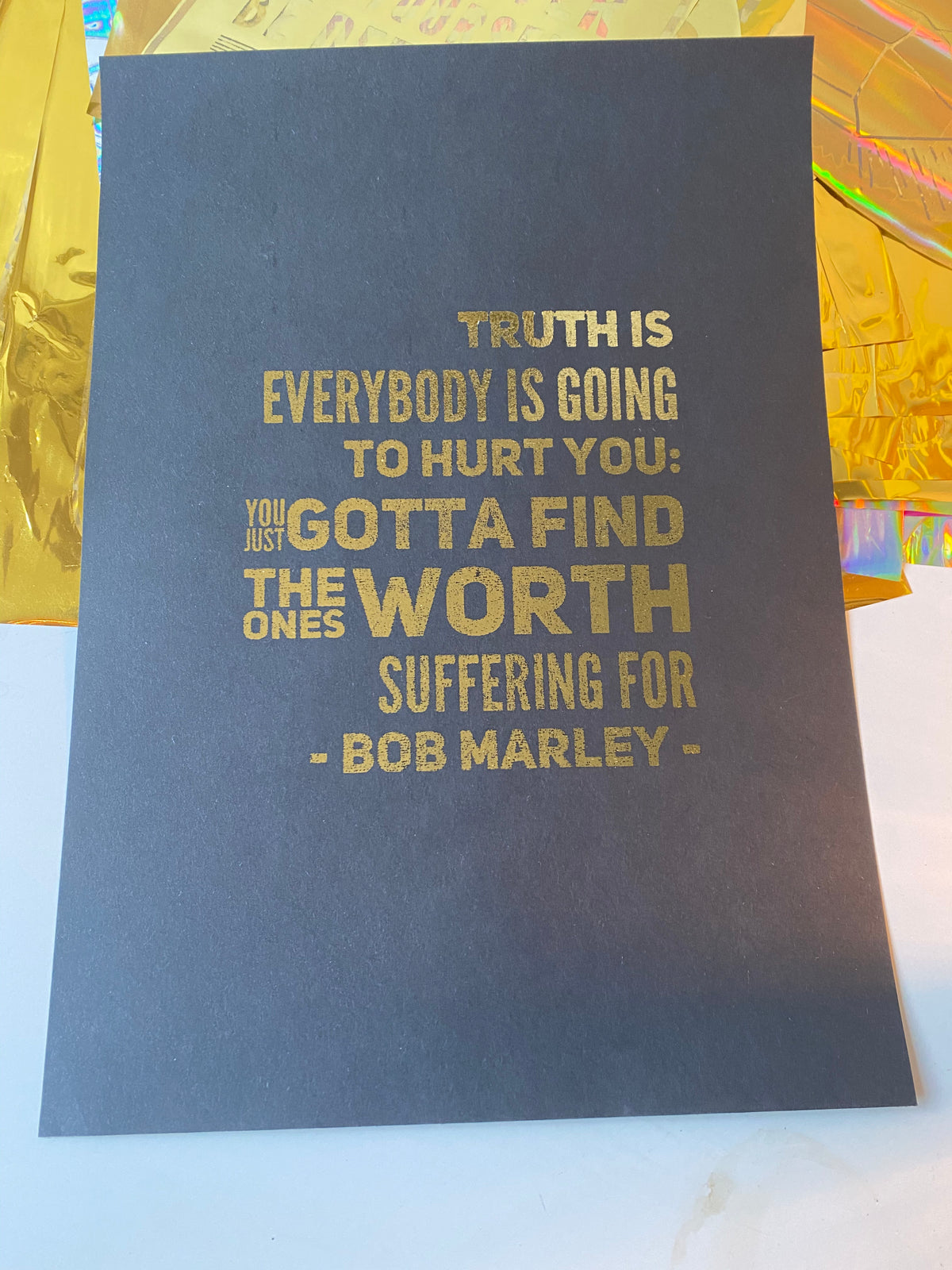 A gold print of a Bob Marley quote about finding the ones worth suffering for, printed in bold gold type on a black background beside iridescent gift wrap - Let's Jam print collection