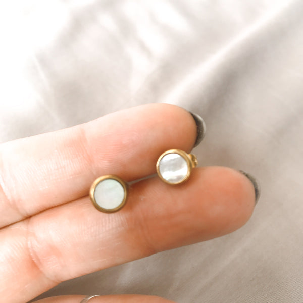 Two round mother-of-pearl stud earrings in a gold-tone stainless steel setting, shown held between fingers to show their soft iridescent shimmer - Let's Jam jewellery collection