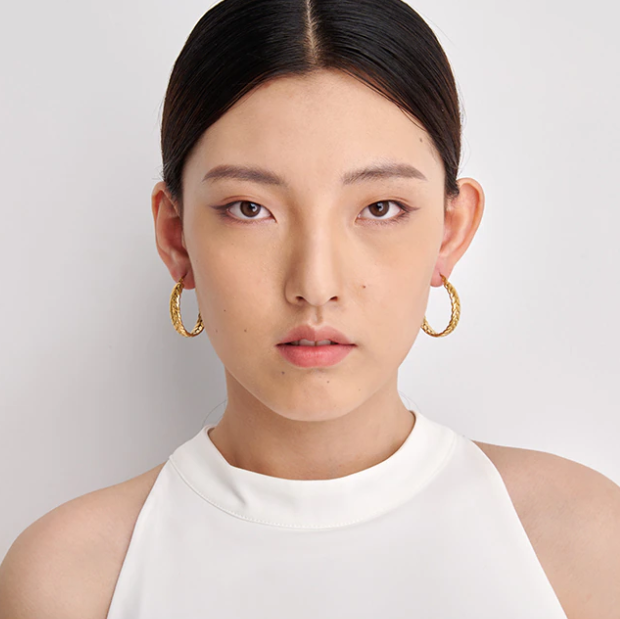 Front-facing portrait of a model wearing chunky gold-plated stainless steel hoop earrings, sleek hair, plain studio background - Let's Jam jewellery collection