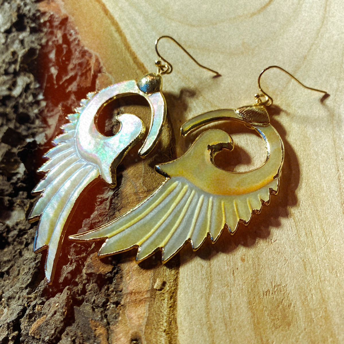 Hand-carved swirled wing-shaped earrings in golden-yellow mother of pearl with gold-plated edging, photographed against natural bark - Let's Jam jewellery collection