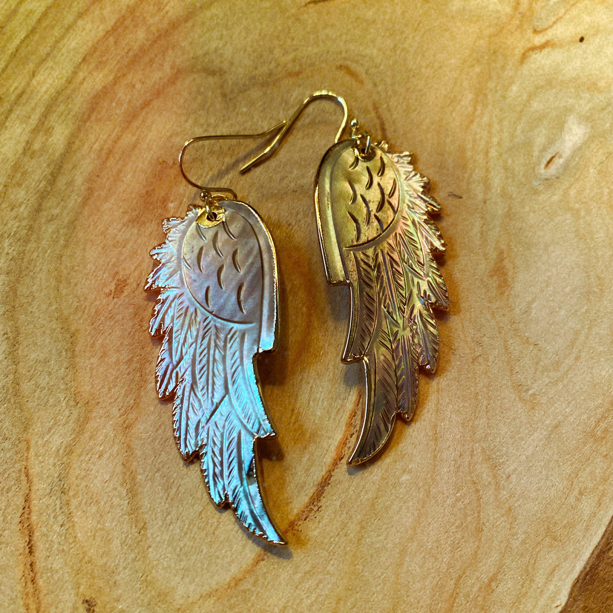 Hand-carved wing-shaped drop earrings in iridescent silver mother of pearl with gold-plated edging, laid on a wooden surface - Let's Jam jewellery collection