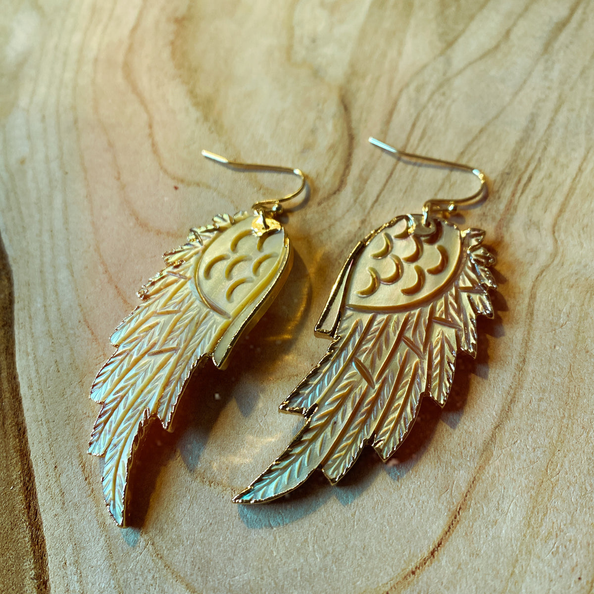 Hand-carved wing-shaped drop earrings in warm golden-yellow mother of pearl with gold-plated edging, laid on a wooden surface - Let's Jam jewellery collection