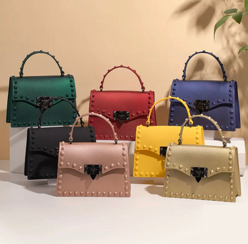 Studded top-handle handbags in green, red, navy, black, pink and yellow with gold rivet detailing, grouped together on a beige surface - Let's Jam collection