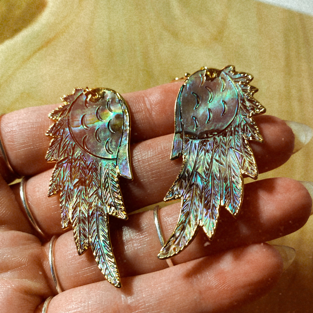 Hand-carved wing-shaped drop earrings in iridescent abalone shell with gold-plated edging, held in hand to show their shimmer - Let's Jam jewellery collection
