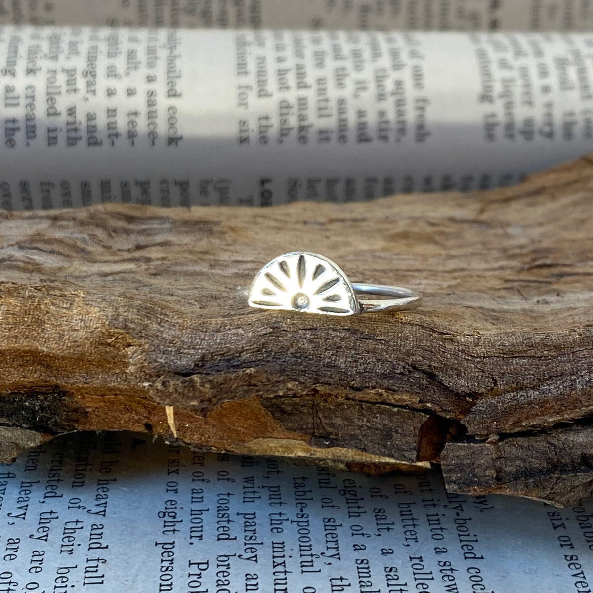 A handcrafted 925 sterling silver ring with a sunburst design, photographed resting on a piece of driftwood beside an open book - Let's Jam jewellery collection