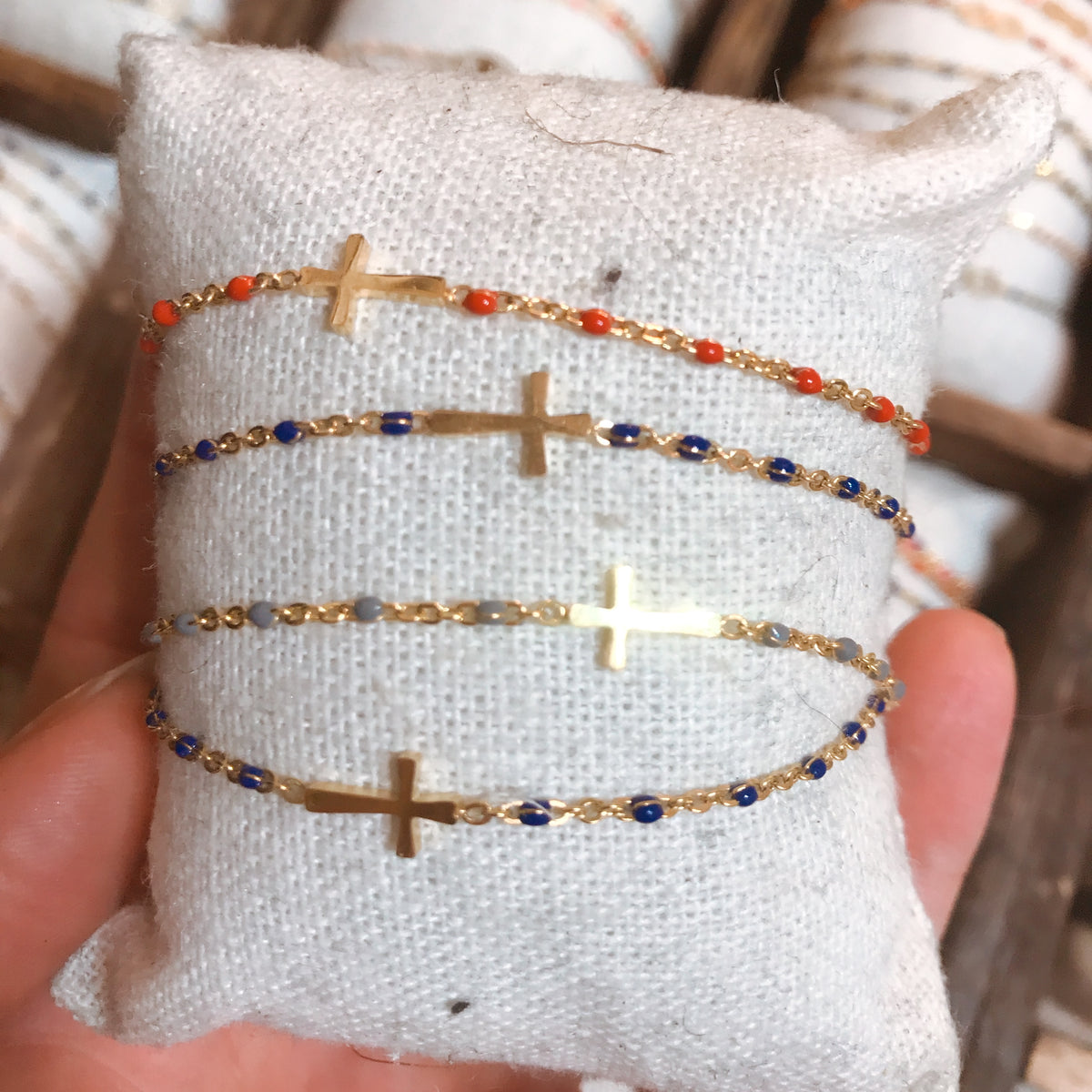 A stack of gold-tone chain bracelets with cross charms and colourful beaded accents, pinned to a small cushion and held in hand - Let's Jam jewellery collection